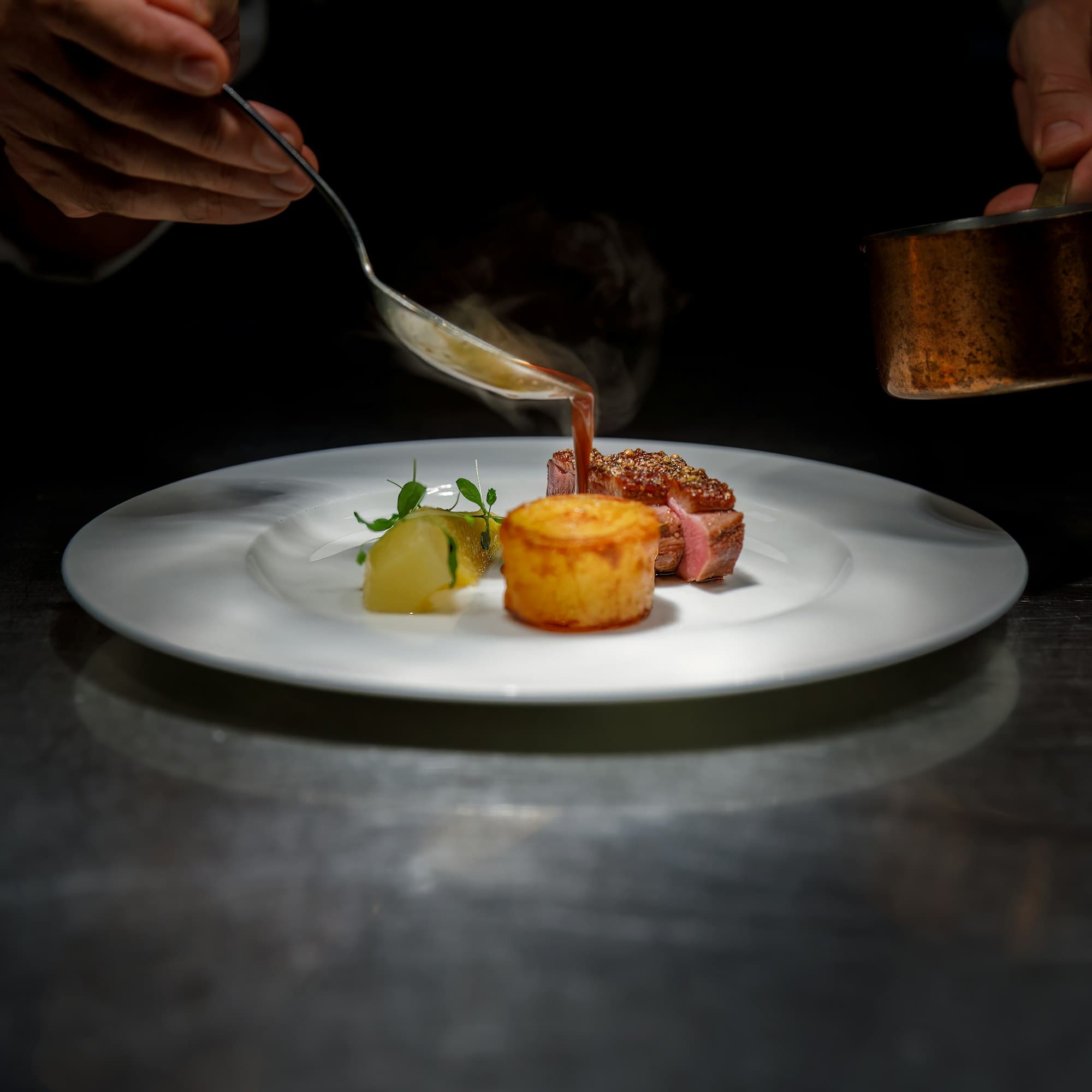 A chef spoons steaming sauce over a plated dish in a dark kitchen