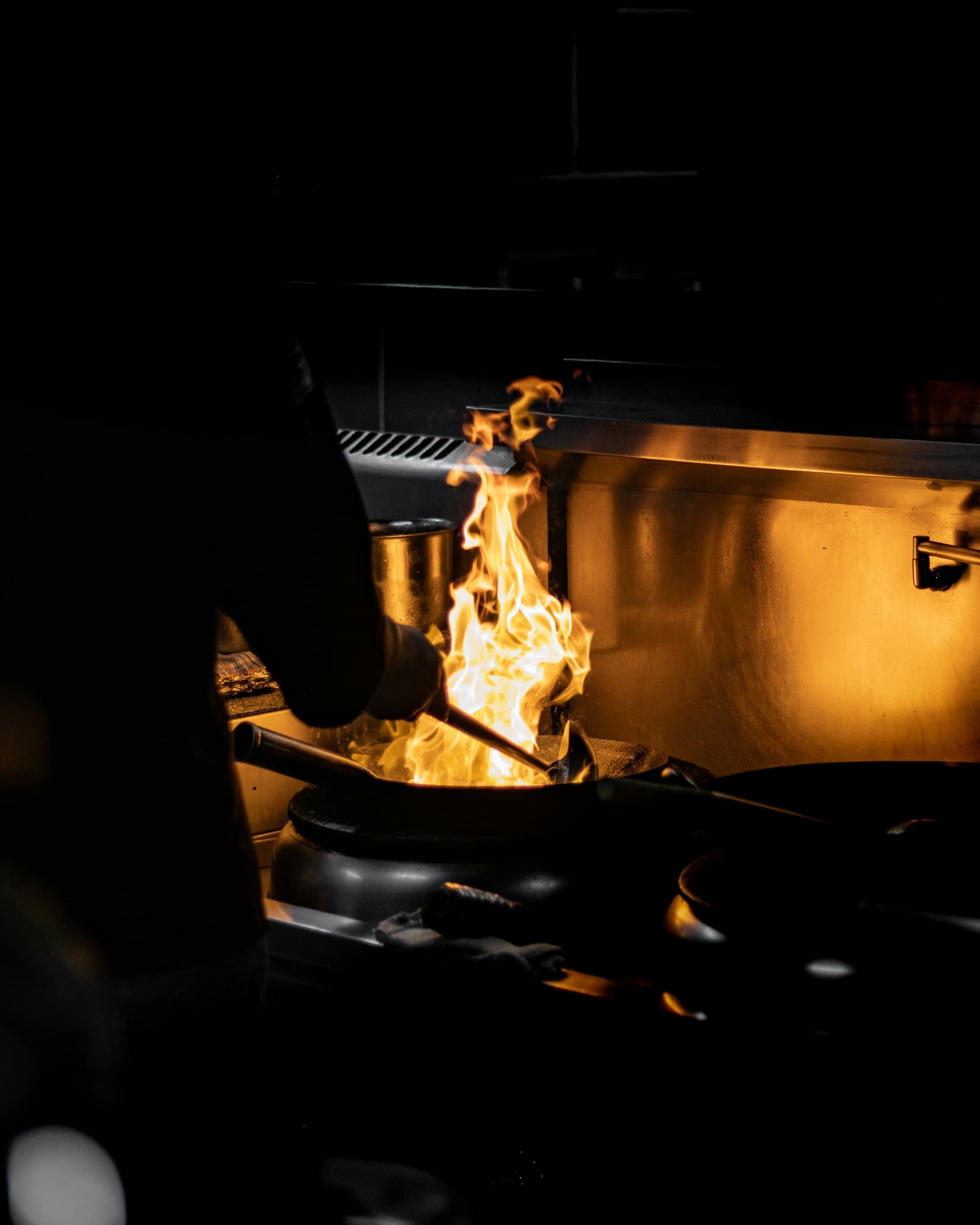 Flames leap from a wok on the stove of a dark commercial kitchen