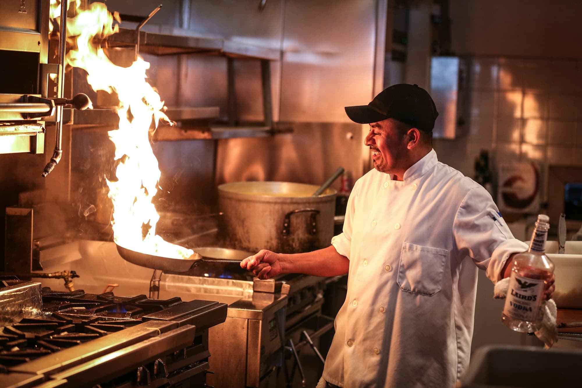 A chef tips a pan into a tall flame on the stove of a dark restaurant kitchen